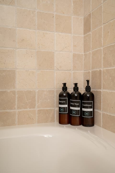 A corner of a bathroom with tiled walls and bottles of shampoo, body wash, and conditioner on the tub edge.
