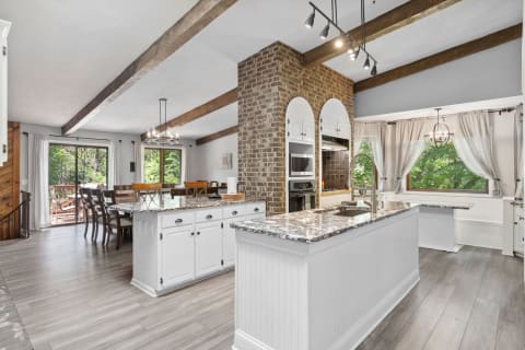 A spacious kitchen featuring a large granite island, wooden beams, and a view of greenery through a window.