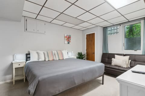 Cozy bedroom with a gray bed and brown sofa, showcasing colorful pillows and natural light.