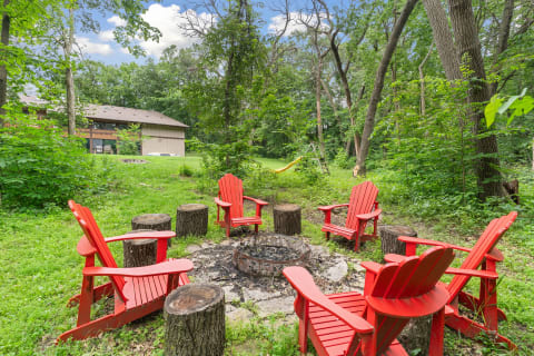 Red Adirondack chairs arranged around a fire pit in a lush green backyard with a cabin in the background.