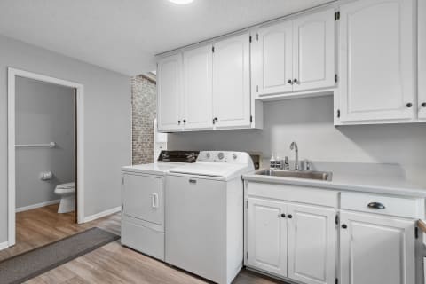 A clean and organized laundry room with a washer, dryer, and sink, showcasing white cabinets and light wood flooring.
