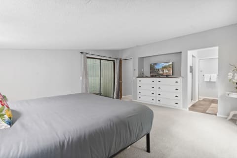 A spacious bedroom with a gray bedspread, white dresser, television, and sliding glass door.