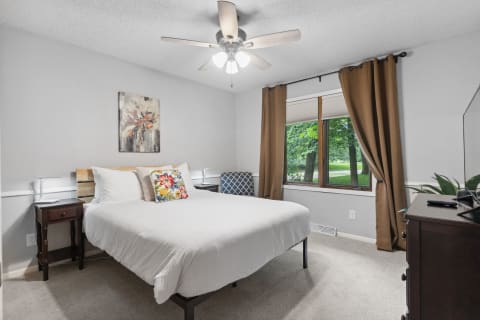 Cozy bedroom with white bed linens, floral pillow, and a large window offering a view of greenery.