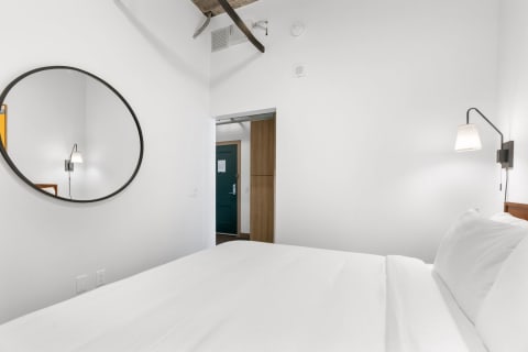 A simple bedroom with white bedding, a circular mirror, and a green door visible in the background.