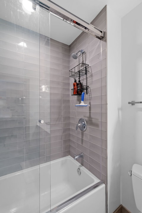 A modern bathroom with a shower enclosed by glass panels, gray tile walls, and a white bathtub.