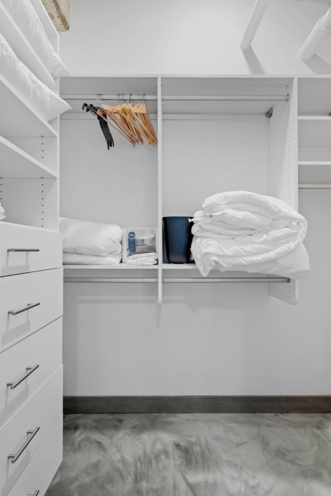 An organized closet with white pillows, wooden hangers, and a black bin on a shelf.