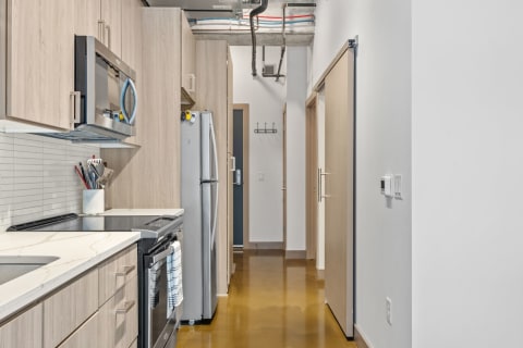 View of a modern kitchen with light wood cabinetry and a hallway leading to another space.