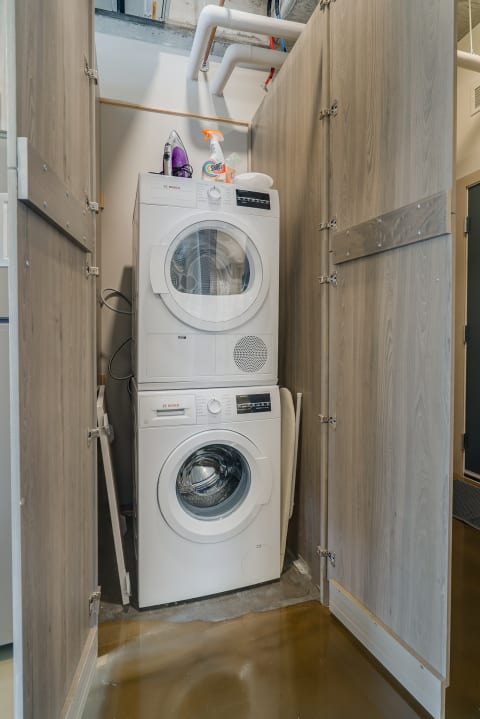 A stacked washer and dryer unit in a compact laundry area, surrounded by wooden cabinets and cleaning supplies.