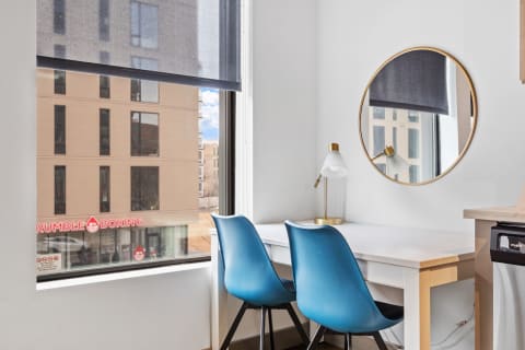 A bright workspace with two blue chairs at a white desk, a lamp, and a mirror, overlooking an urban view.