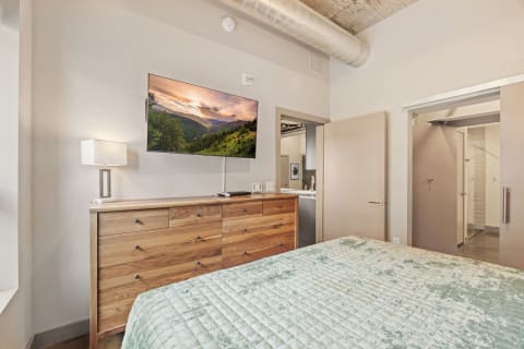 A contemporary bedroom with a wooden dresser, a large TV, and a mint green quilt on the bed.