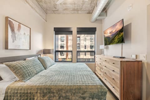 Cozy modern bedroom featuring a quilted bedspread, abstract artwork, and large windows.