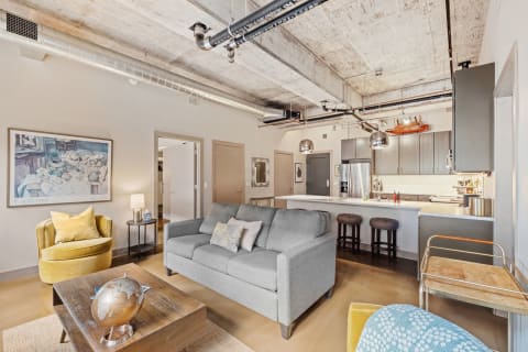 Inside view of a contemporary loft apartment featuring a living area with a gray sofa, yellow armchair, and a kitchen in the background.