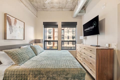 Modern bedroom with green quilted bed and urban views through large windows.