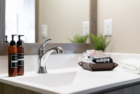 Modern bathroom sink with chrome faucet, amber lotion and soap bottles, and makeup supplies in a brown container.