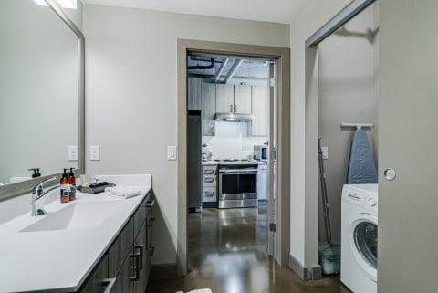 Modern bathroom with sleek design, featuring a sink, mirror, and doorway to a kitchen and laundry nook.
