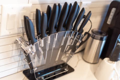 Knife block with an array of black-handled kitchen knives against a white tiled wall.