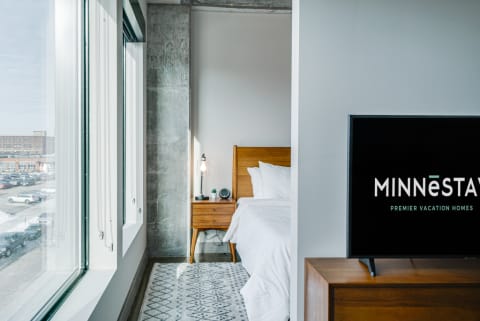 Stylish bedroom interior featuring large windows, a cozy bed, and a TV with a vacation rental logo.