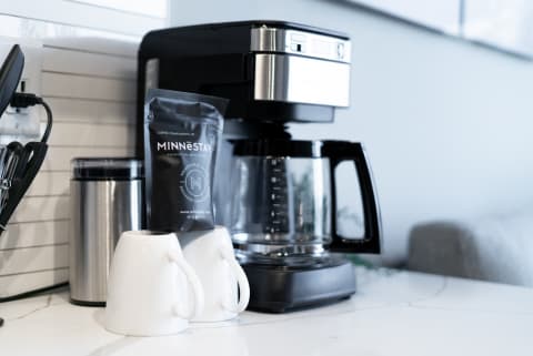 A coffee maker with a coffee package and two white mugs on a marble countertop.