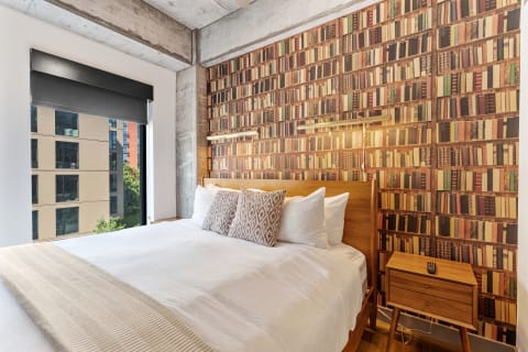 A modern bedroom featuring a bed with white linens, book-patterned wallpaper, and a view of an urban building.