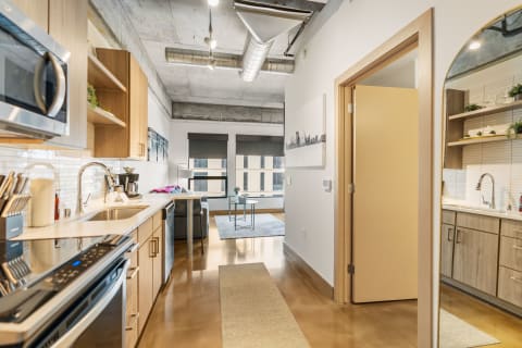 Modern kitchen with wooden cabinets and stainless steel appliances, leading to a living room area.