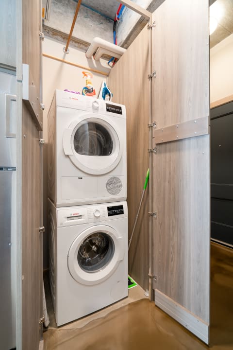 Compact space with a washer and dryer stacked between wooden panels and cleaning supplies on a shelf.