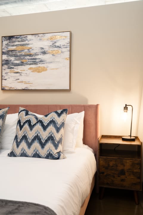 Cozy bedroom corner with a pink bed frame, patterned pillows, abstract wall art, and a wooden nightstand with a lamp.