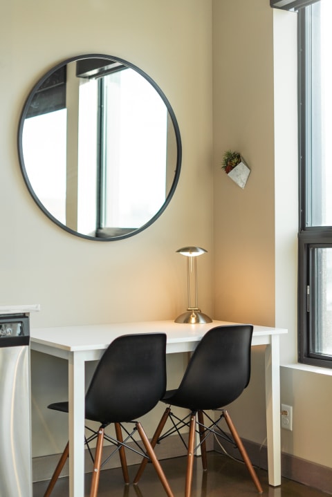 A minimalist workspace with a white desk, black chairs, and a round mirror, illuminated by a lamp.