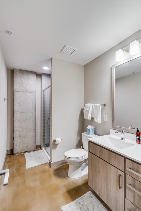 A contemporary bathroom with a light wood vanity, white sink, toilet, and shower surrounded by a concrete wall.
