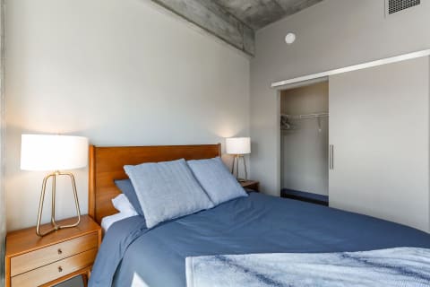 Modern bedroom with a blue bedspread, wooden furniture, and an industrial ceiling.