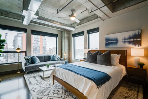 A stylish bedroom with a large bed, gray sofa, and modern decor, featuring large windows and an urban view.