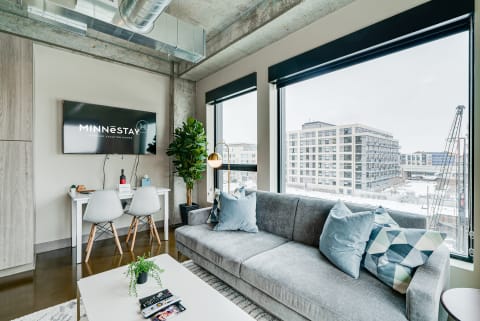 A stylish modern living room featuring a gray sofa, white desk, and large windows with urban views.