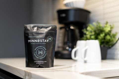 A coffee package from MINNēSTAY on a marble countertop next to a coffee maker and white mugs.