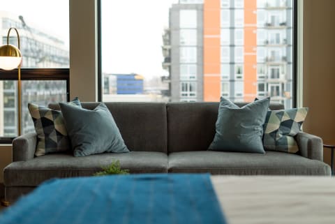 A stylish living room featuring a grey sofa with blue and patterned pillows, viewed through a large window.