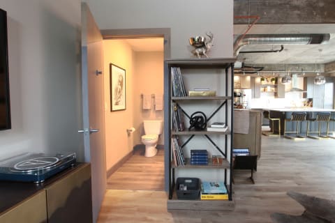 Interior view of a modern apartment with a bookshelf, bathroom doorway, and kitchen in the background.