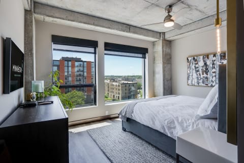 Modern bedroom with large windows, a gray bed, and abstract wall art overlooking the city skyline.