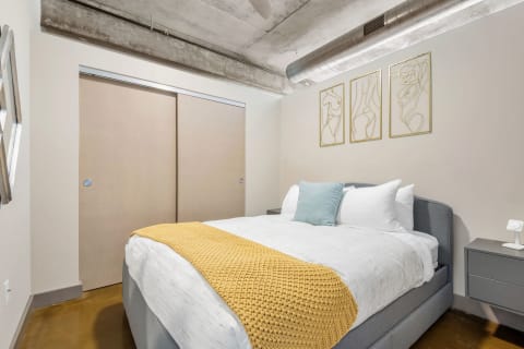 A cozy bedroom featuring a bed with white linens, yellow throw blanket, and abstract wall art.