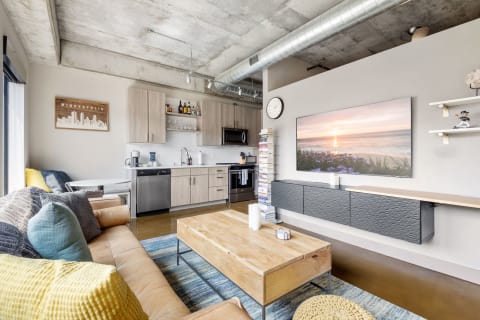 A modern living room showcasing a kitchen, cozy sofa, wooden coffee table, and large screen TV, all in an industrial design.