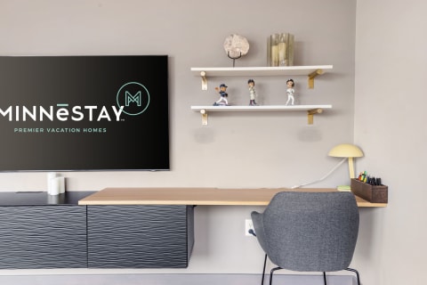 A contemporary workspace with a desk, chair, and shelves displaying figurines, alongside a MINNĒSTAY logo on a wall-mounted screen.
