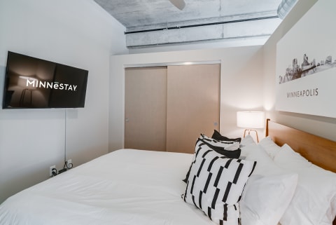Modern bedroom featuring a bed with decorative pillows and a TV displaying MINNēSTAY.
