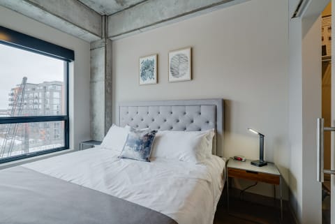 Modern bedroom with gray headboard, decorative pillow, and urban view outside the window.