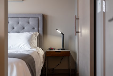 A bedroom with a grey tufted headboard, white bedding, a black desk lamp, and an alarm clock showing 1:09.