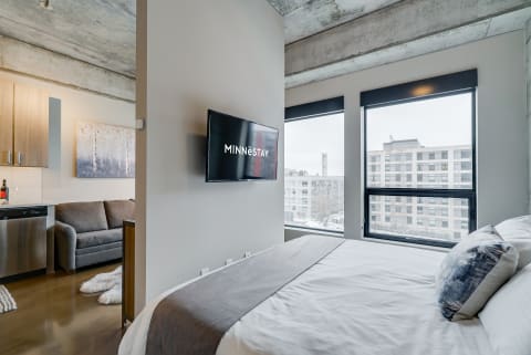 Interior of a modern hotel room with a bed, kitchenette, and large windows.