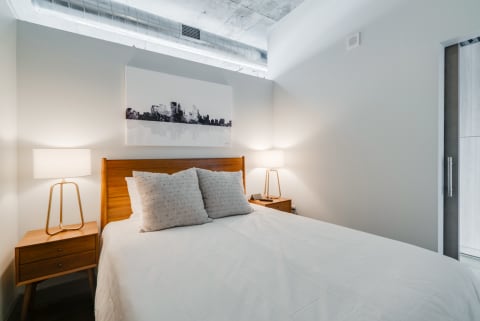 Modern bedroom with a white bed, wooden nightstands, lamps, and skyline artwork on the wall.
