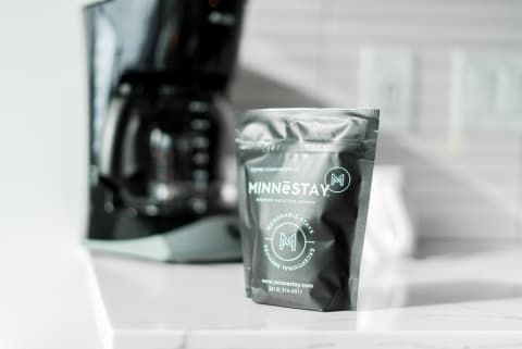 Coffee packet labeled MINNĒSTAY in front of a modern coffee maker.