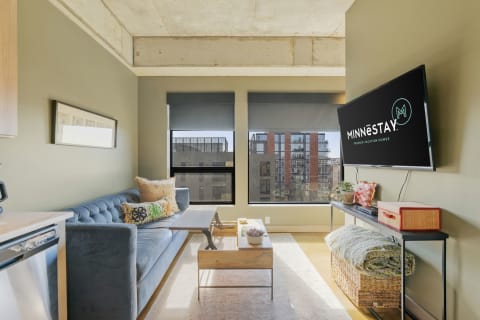 Modern living room with blue sofa, large windows, and a 'Minnestay' TV.