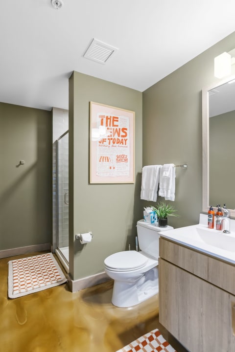 A contemporary bathroom featuring olive green walls, a glass shower, and light wood vanity with a pop of color from a poster and towels.