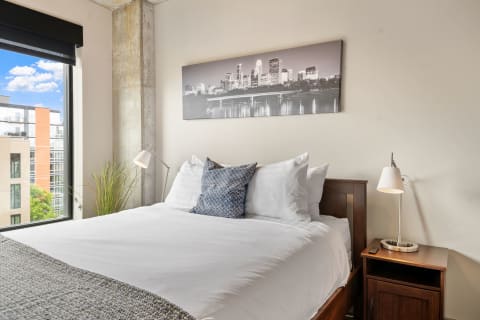 A cozy bedroom with a double bed, plush pillows, a modern lamp, and a city skyline print above the bed.