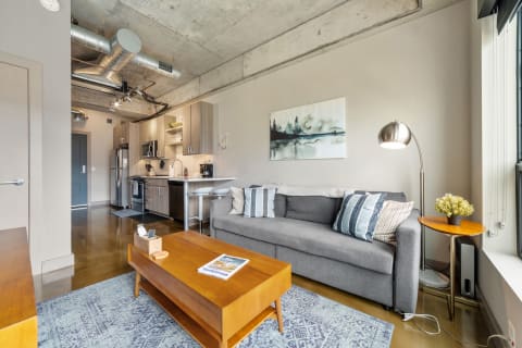 Interior view of a modern studio apartment featuring a gray couch, wooden coffee table, and kitchen space.