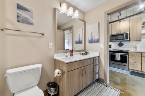 Contemporary bathroom with white toilet, wooden vanity, and a view of the kitchen.