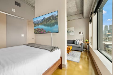 Interior of a modern studio apartment with a bed, mounted TV, sofa, and city view.